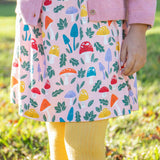 A child standing on grass and wearing the Frugi kids pelican fun-guys amelia dress. A GOTS organic cotton dress in pink with multiple playful woodland inspired prints. 