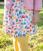 A child standing on grass and wearing the Frugi kids pelican fun-guys amelia dress. A GOTS organic cotton dress in pink with multiple playful woodland inspired prints. 
