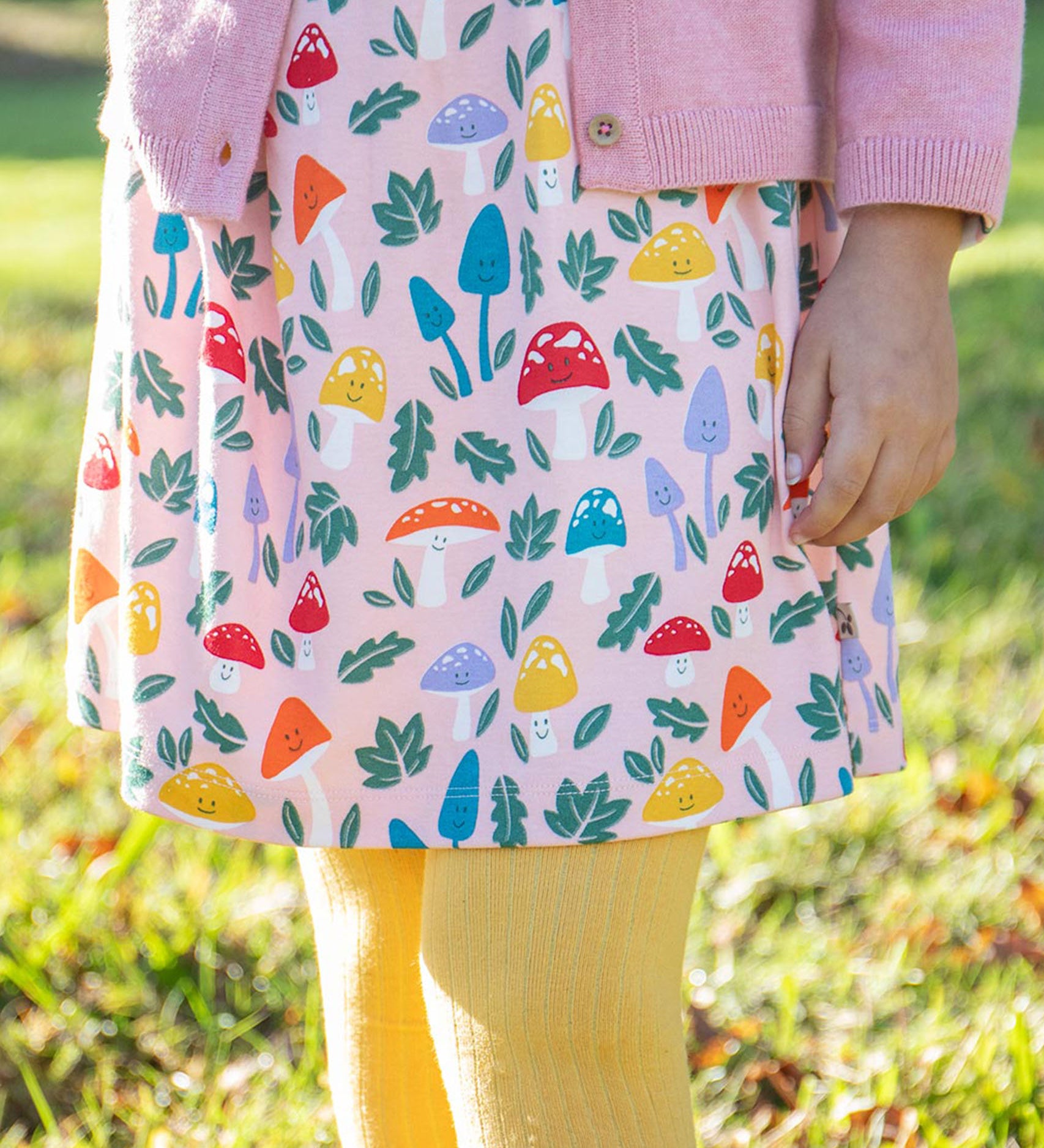 A child standing on grass and wearing the Frugi kids pelican fun-guys amelia dress. A GOTS organic cotton dress in pink with multiple playful woodland inspired prints. 