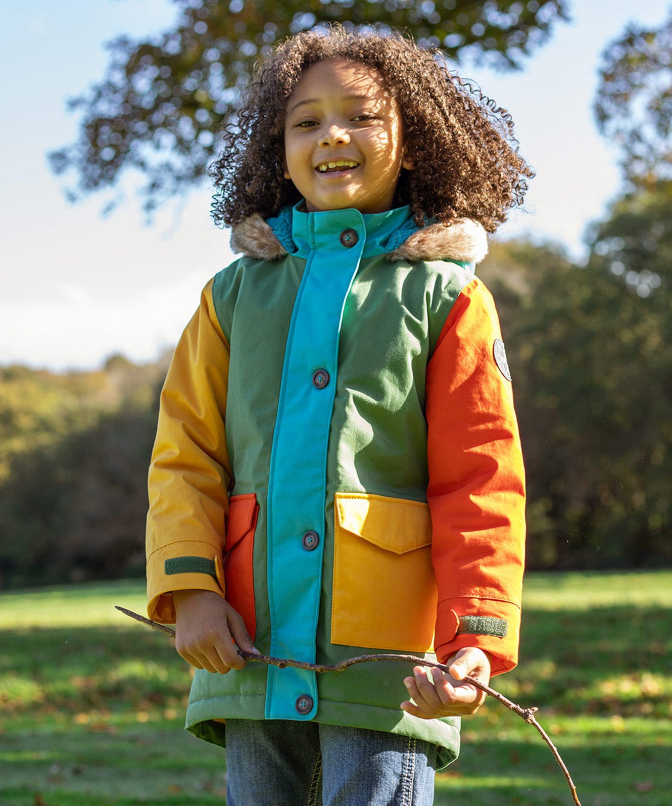 A child wearing the Frugi kids penhale Khaki parka outside- A long length thick warm coat  furry hood for warmth and  dark orange and yellow side pockets.