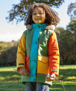 A child wearing the Frugi kids penhale Khaki parka outside- A long length thick warm coat  furry hood for warmth and  dark orange and yellow side pockets.
