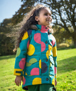 A child wearing the Frugi kids penhale super spot parka outside- A long length  organic cotton fabric warm coat with furry hood for warmth. Features a velcro strap on the arms to adjust the fit  