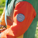 A close up of the orange arm of the Frugi penhale khaki colour block parka showing the circular brand patch stitched on the top of the arm on organic cotton fabric