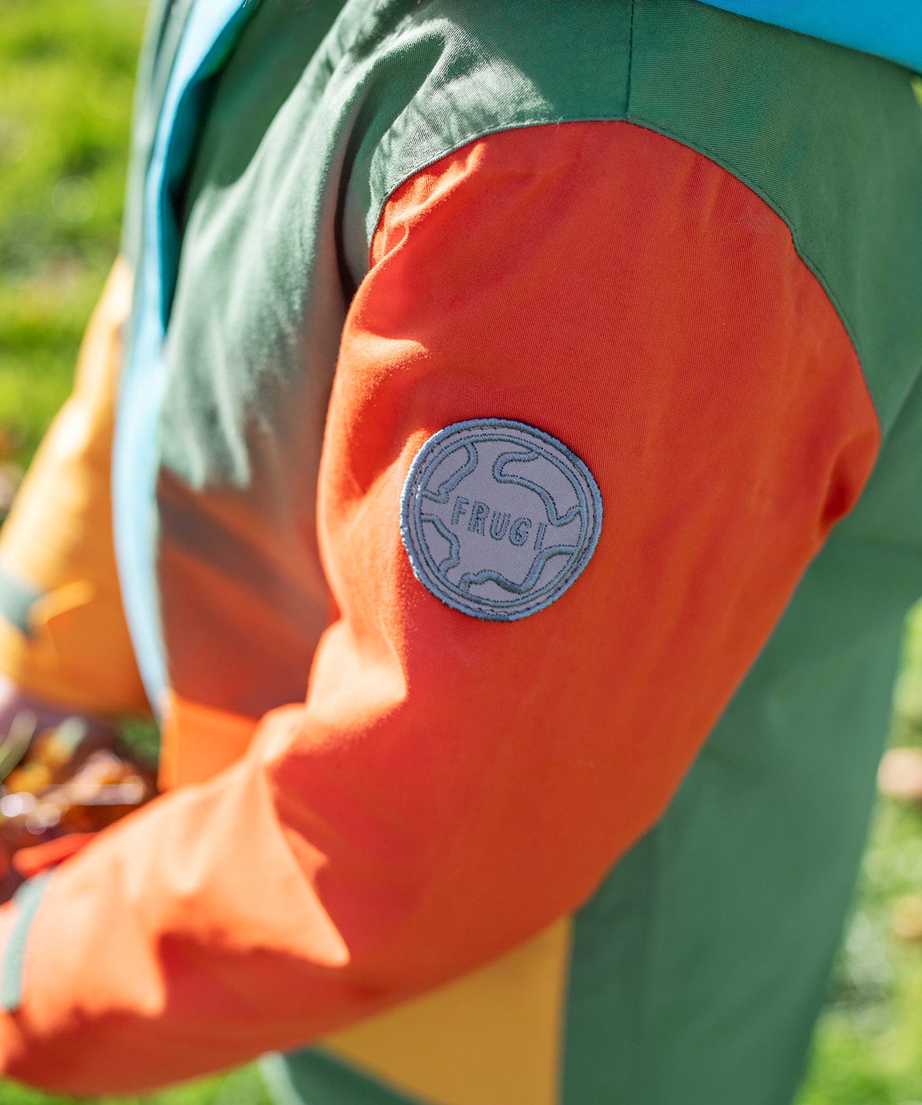 A close up of the orange arm of the Frugi penhale khaki colour block parka showing the circular brand patch stitched on the top of the arm on organic cotton fabric