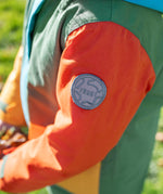 A close up of the orange arm of the Frugi penhale khaki colour block parka showing the circular brand patch stitched on the top of the arm on organic cotton fabric