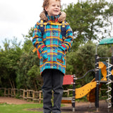 Child wearing Frugi's Penhale parka coat in a blue red and yellow tartan check with the faux fur trimmed hood down standing on a blue table in a playground.