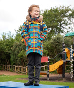 Child wearing Frugi's Penhale parka coat in a blue red and yellow tartan check with the faux fur trimmed hood down standing on a blue table in a playground.