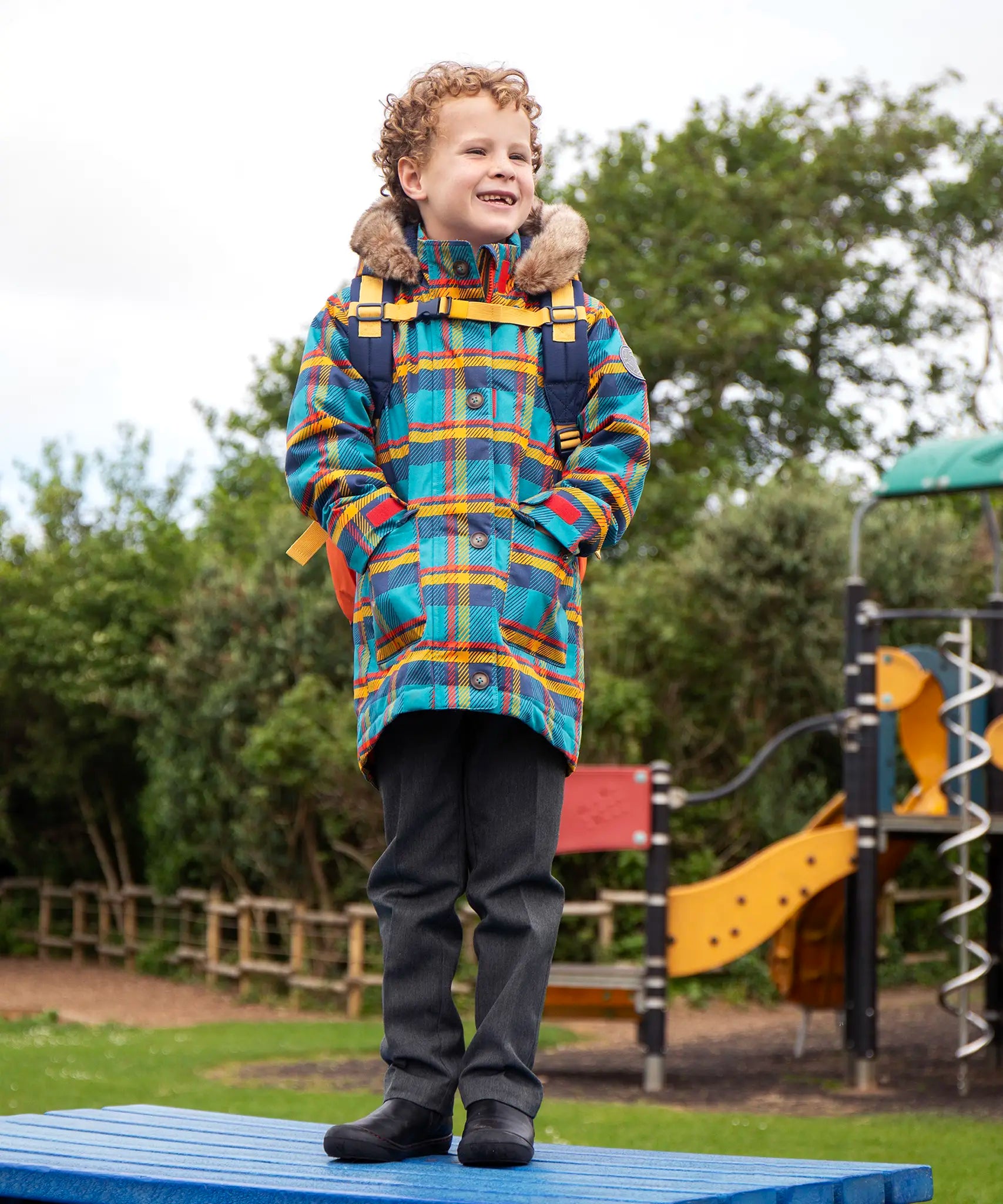 Child wearing Frugi's Penhale parka coat in a blue red and yellow tartan check with the faux fur trimmed hood down standing on a blue table in a playground.