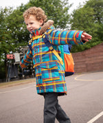 Child wearing Frugi's Penhale parka coat in a blue red and yellow tartan check with the faux fur trimmed hood down in a playground.