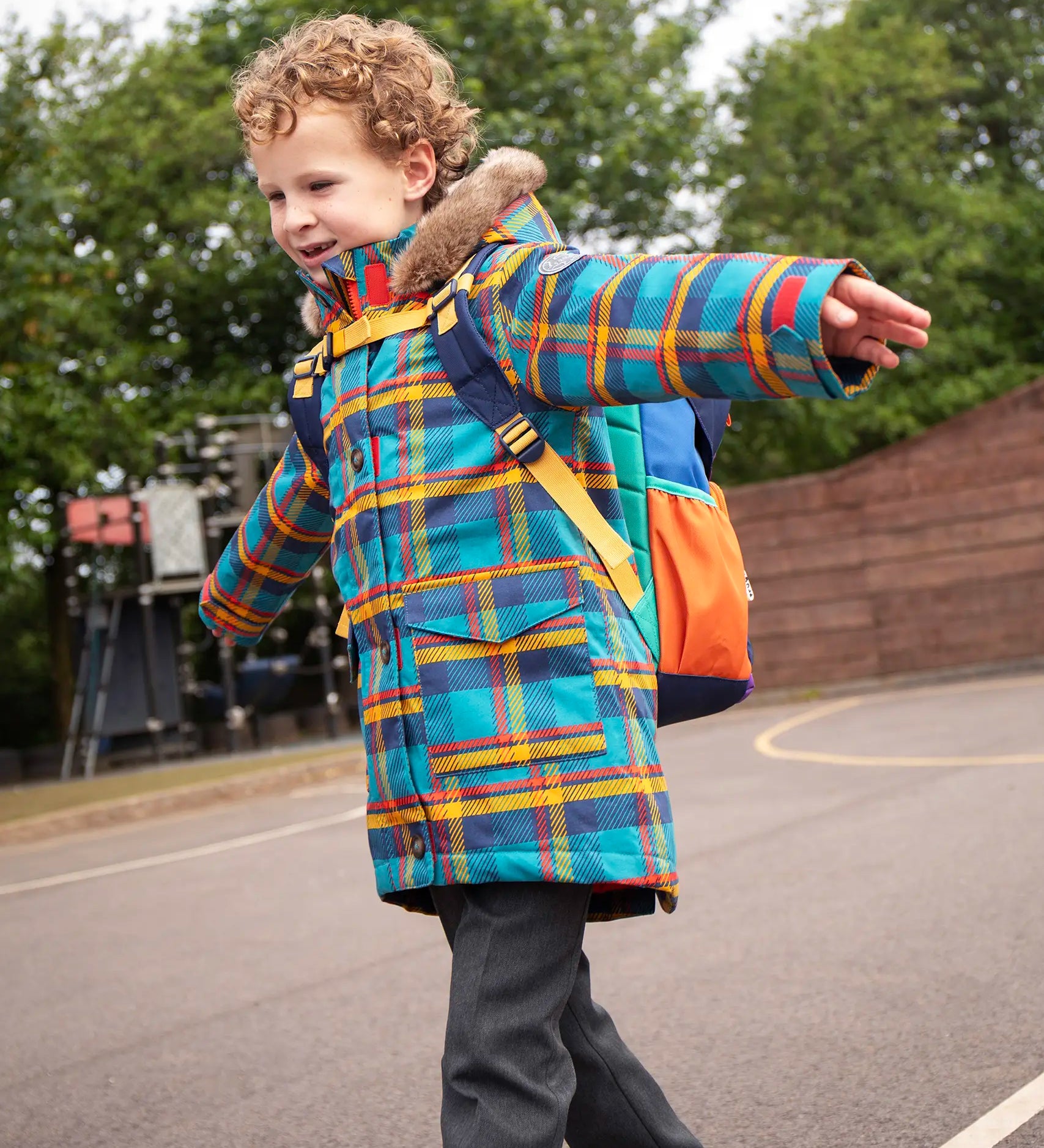 Child wearing Frugi's Penhale parka coat in a blue red and yellow tartan check with the faux fur trimmed hood down in a playground.
