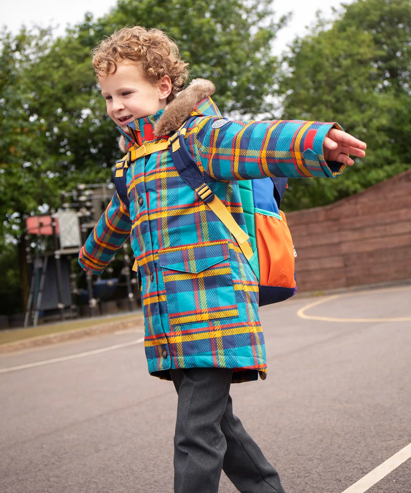 Child wearing Frugi's Penhale parka coat in a blue red and yellow tartan check with the faux fur trimmed hood down in a playground.