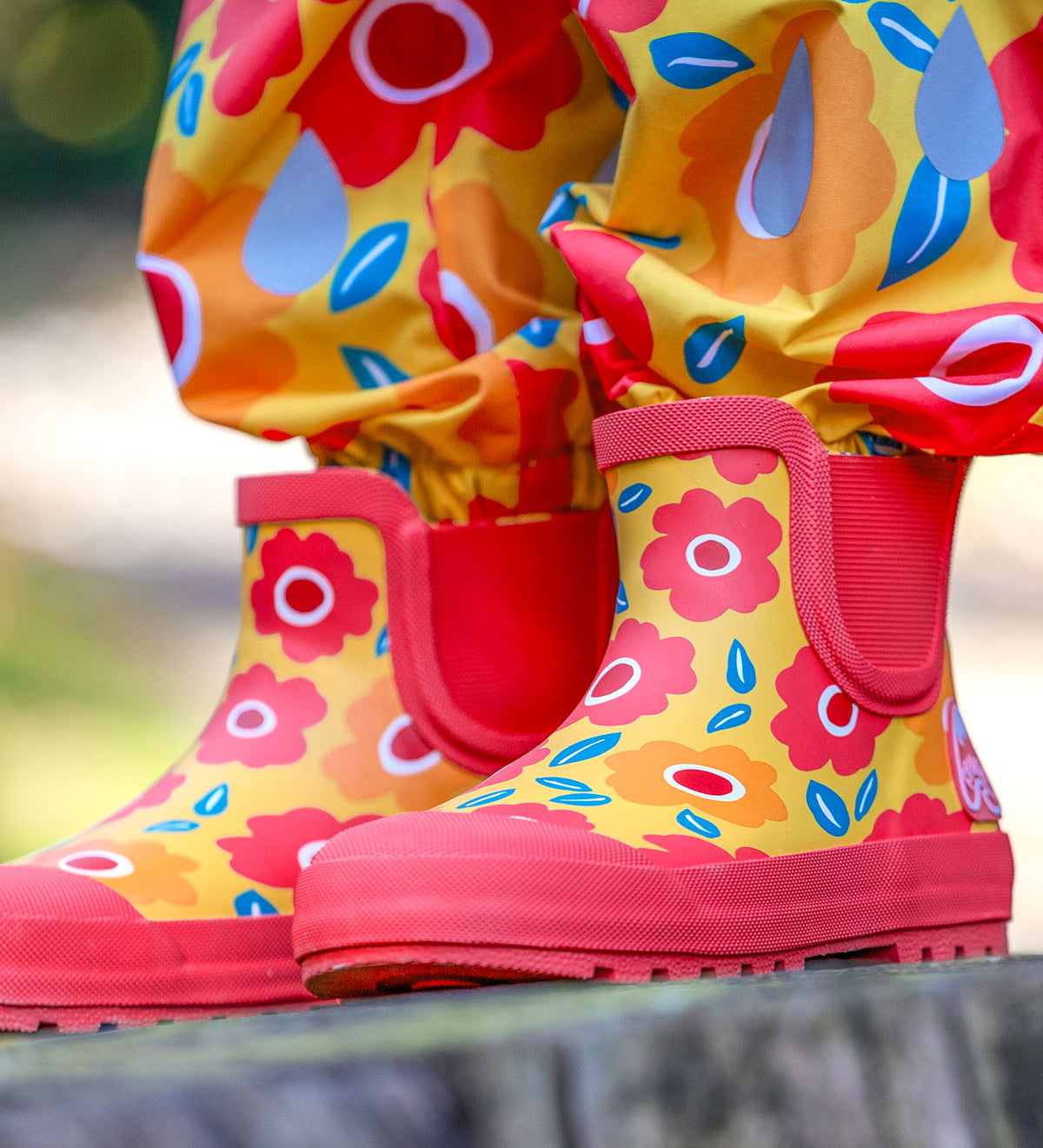 A close up of a child's feet wearing the Frugi rain or shine short welly boots with a floral fields design. A yellow background  with orange and pink  flower print  and pink soles and binding. They are also wearing matching waterproof rain trousers. 