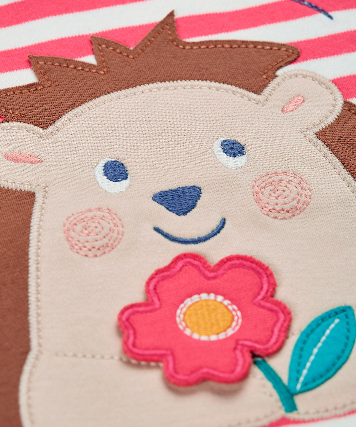 A close up of the hedgehog applique on the front of the Frugi Easy On pink breton stripe hedgehog applique short sleeved organic cotton T-Shirt. The hedgehog is holding a pink flower and there is a little bee flying above.