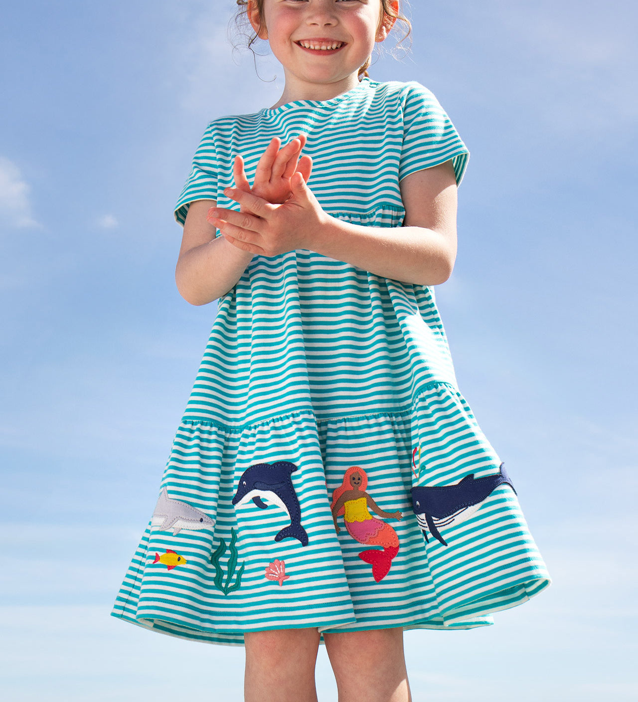 A smiling young child wearing the Frugi Pixy Sea Moss Mini Stripe Dress, with a Sealife print along the bottom, featuring whales, dolphins, sharks, a Mermaid and some small fish, surrounded by colorful seaweed, stood outside.