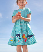 A smiling young child wearing the Frugi Pixy Sea Moss Mini Stripe Dress, with a Sealife print along the bottom, featuring whales, dolphins, sharks, a Mermaid and some small fish, surrounded by colorful seaweed, stood outside.