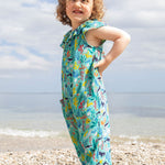 A smiling toddler wearing the Frugi Maris Playsuit, featuring a Sea Life print which includes different sea animals and colourful seaweed and coral, standing on a pebbled beach.