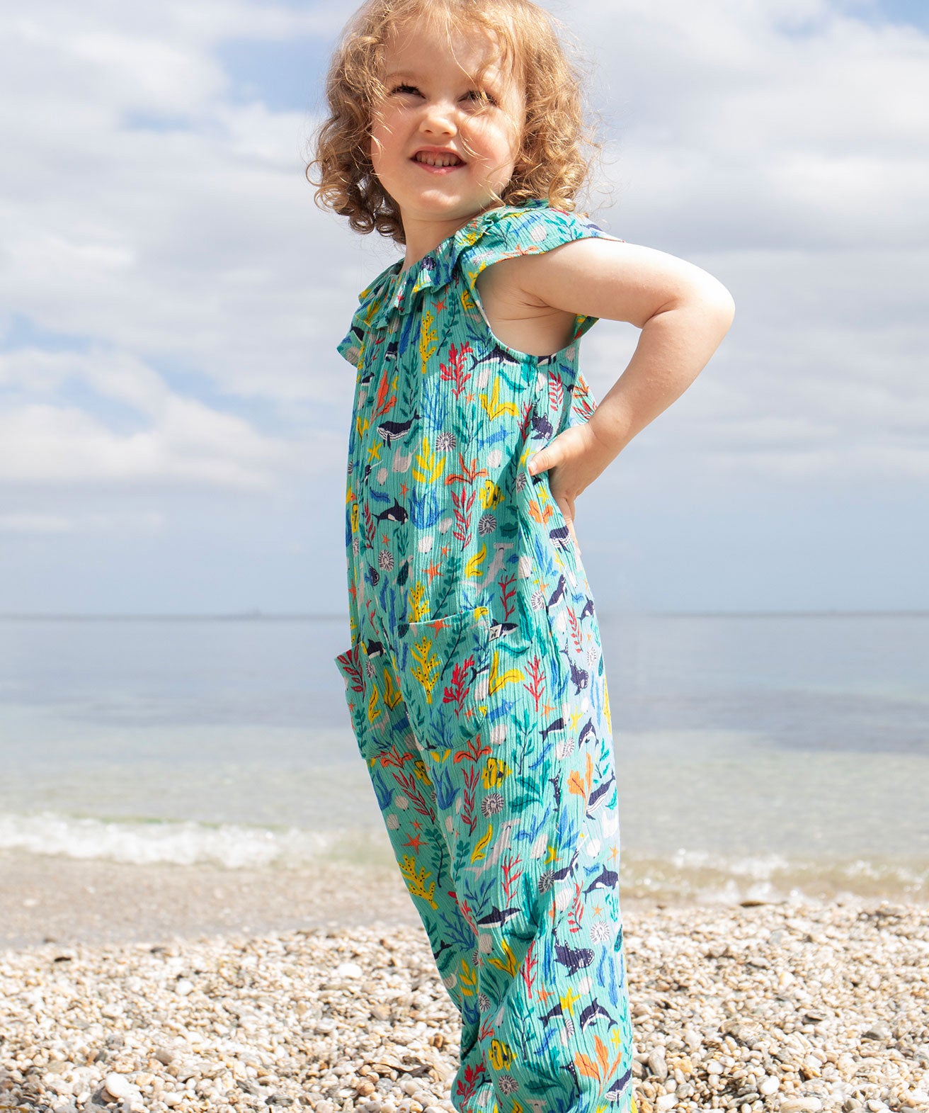 A smiling toddler wearing the Frugi Maris Playsuit, featuring a Sea Life print which includes different sea animals and colourful seaweed and coral, standing on a pebbled beach.