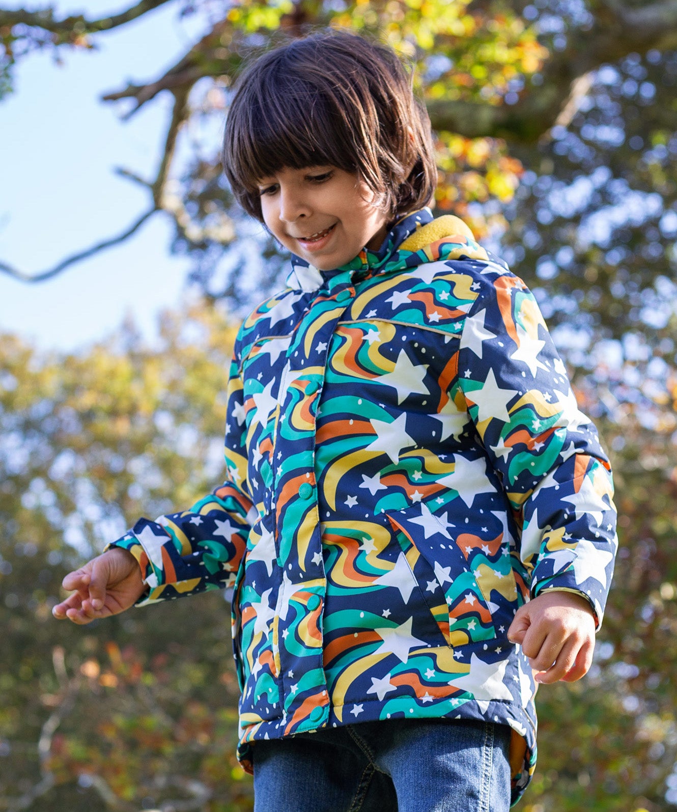 A toddler standing outside wearing the Frugi playtime magic stars coat. A thick long length hooded coat with a playful outer space inspired design