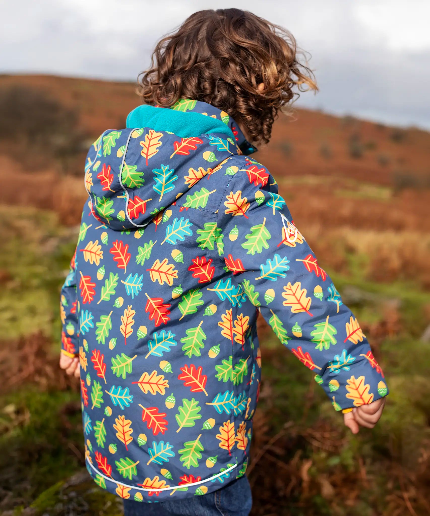 Back of small child wearing the Frugi playtime waterproof coat in navy with a repeating multicoloured acorn design. Showing the hood is down. 