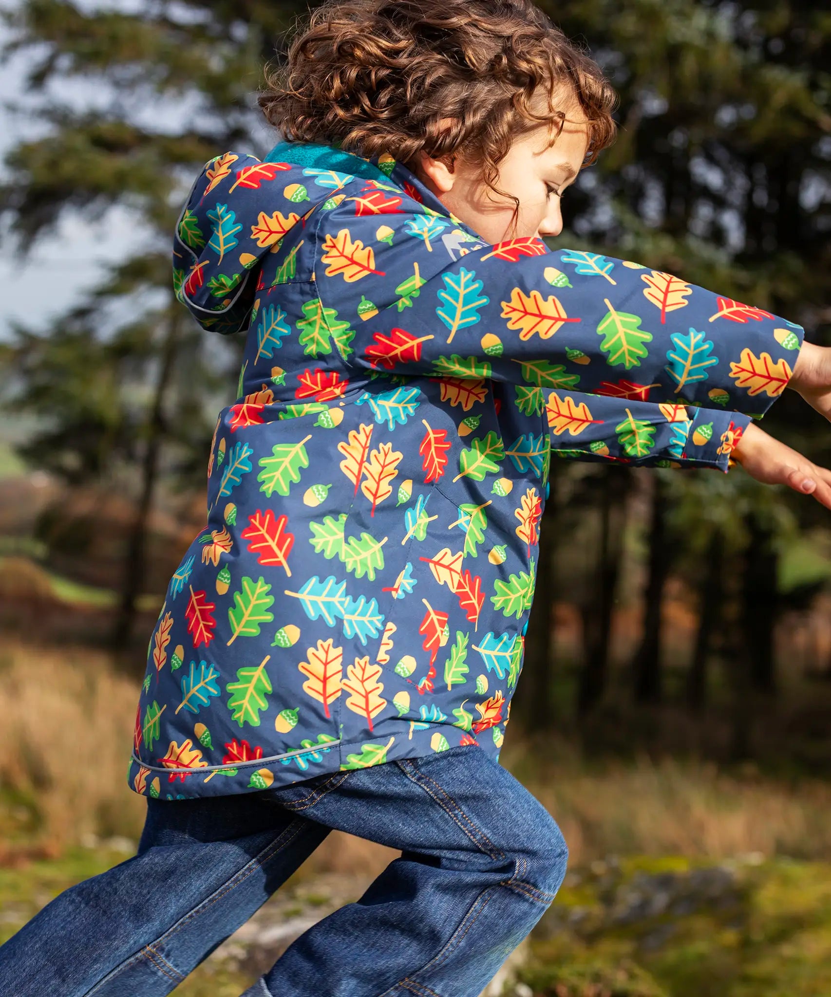 side of small child wearing the Frugi playtime waterproof coat in navy with a repeating multicoloured acorn design. Coat has a relaxed fit