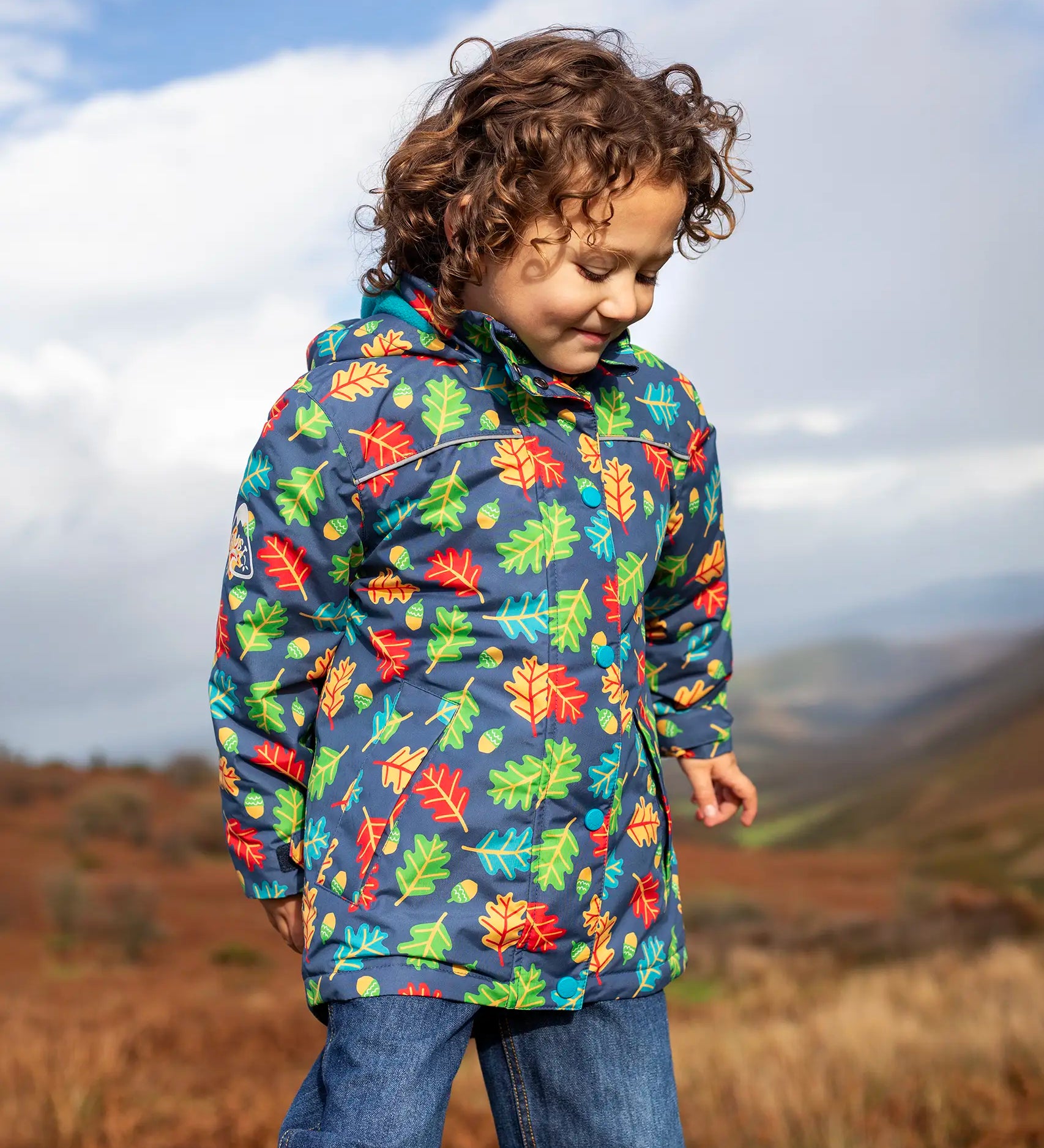 Small child wearing the Frugi playtime waterproof coat in navy with a repeating multicoloured acorn design. Showing the hood is down. 