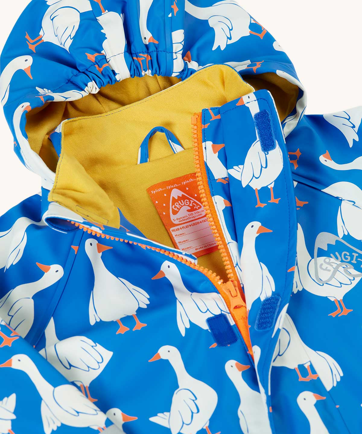 A close up of the top of the orange zip opening on the Frugi cobalt blue rain jacket with all over goose print. The jacket has a yellow jersey lining and a label underneath the hanging loop inside with space to write names. 