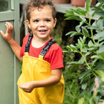 A little boy wearing the Frugi Bumble Bee Yellow waterproof recycled Puddle Buster trousers in a garden shed