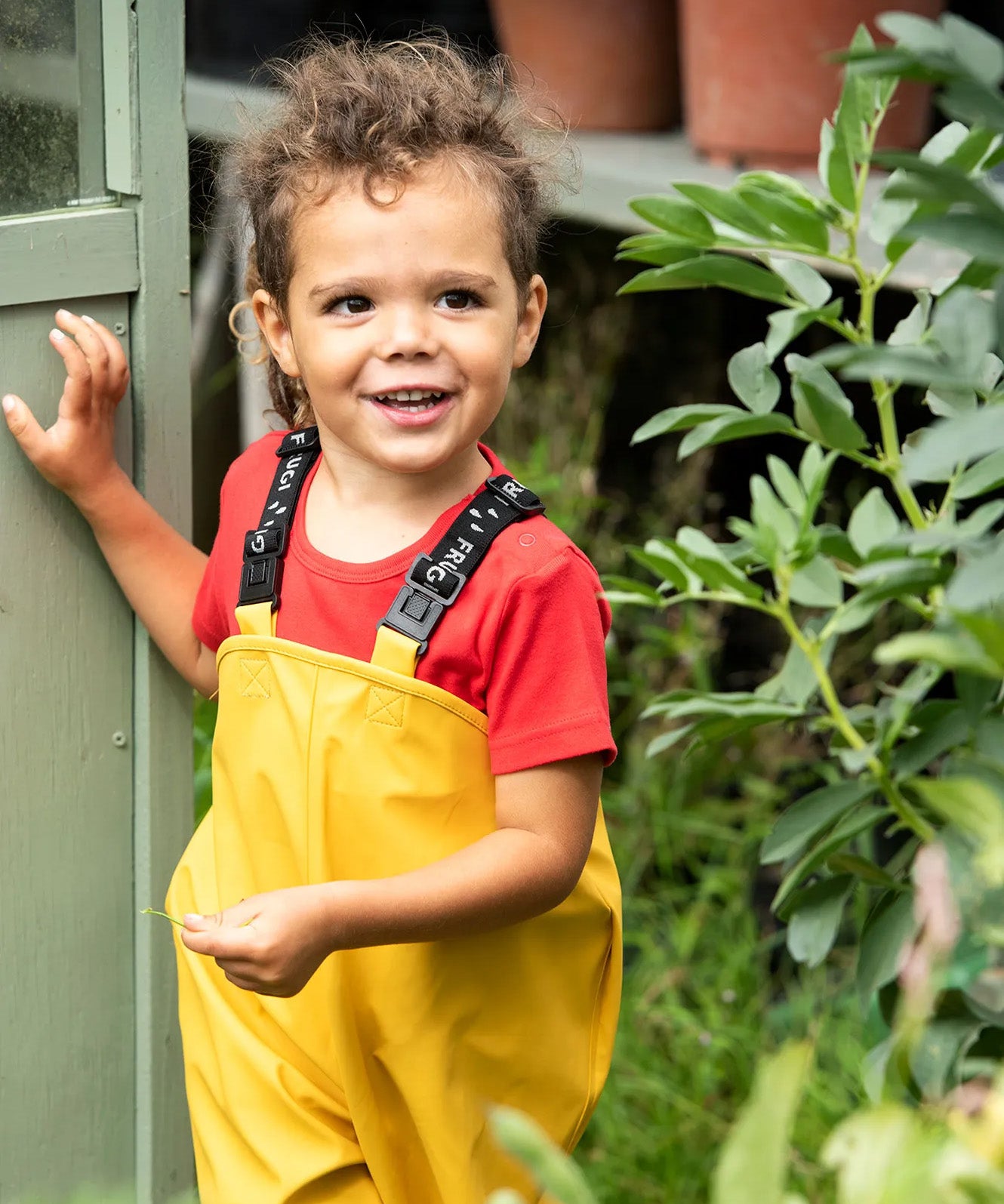 A little boy wearing the Frugi Bumble Bee Yellow waterproof recycled Puddle Buster trousers in a garden shed