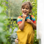 A little girl wearing Frugi waterproof Puddle Buster trousers in a garden, they're made from recycled plastic bottles and come in bright yellow