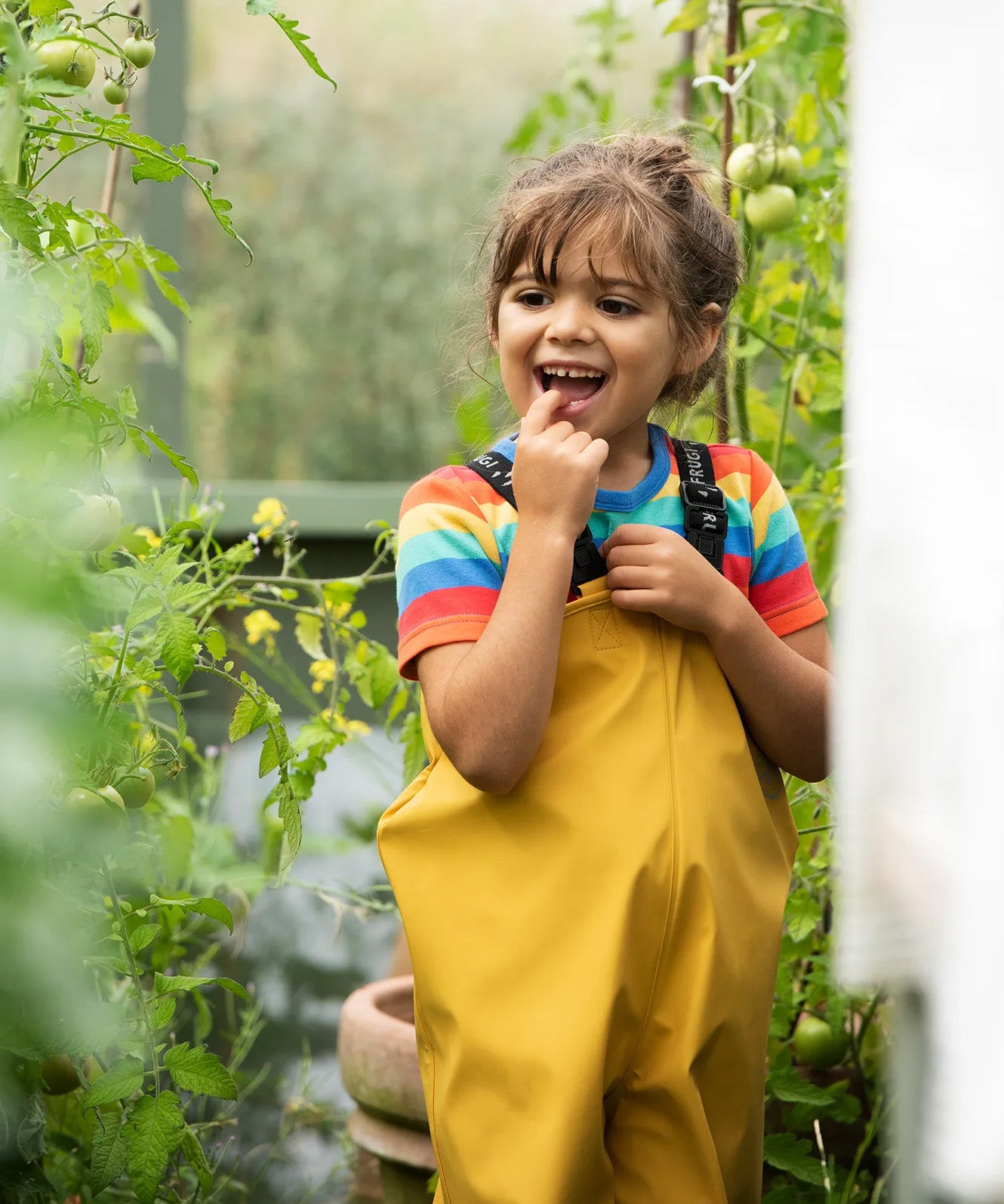 A little girl wearing Frugi waterproof Puddle Buster trousers in a garden, they're made from recycled plastic bottles and come in bright yellow