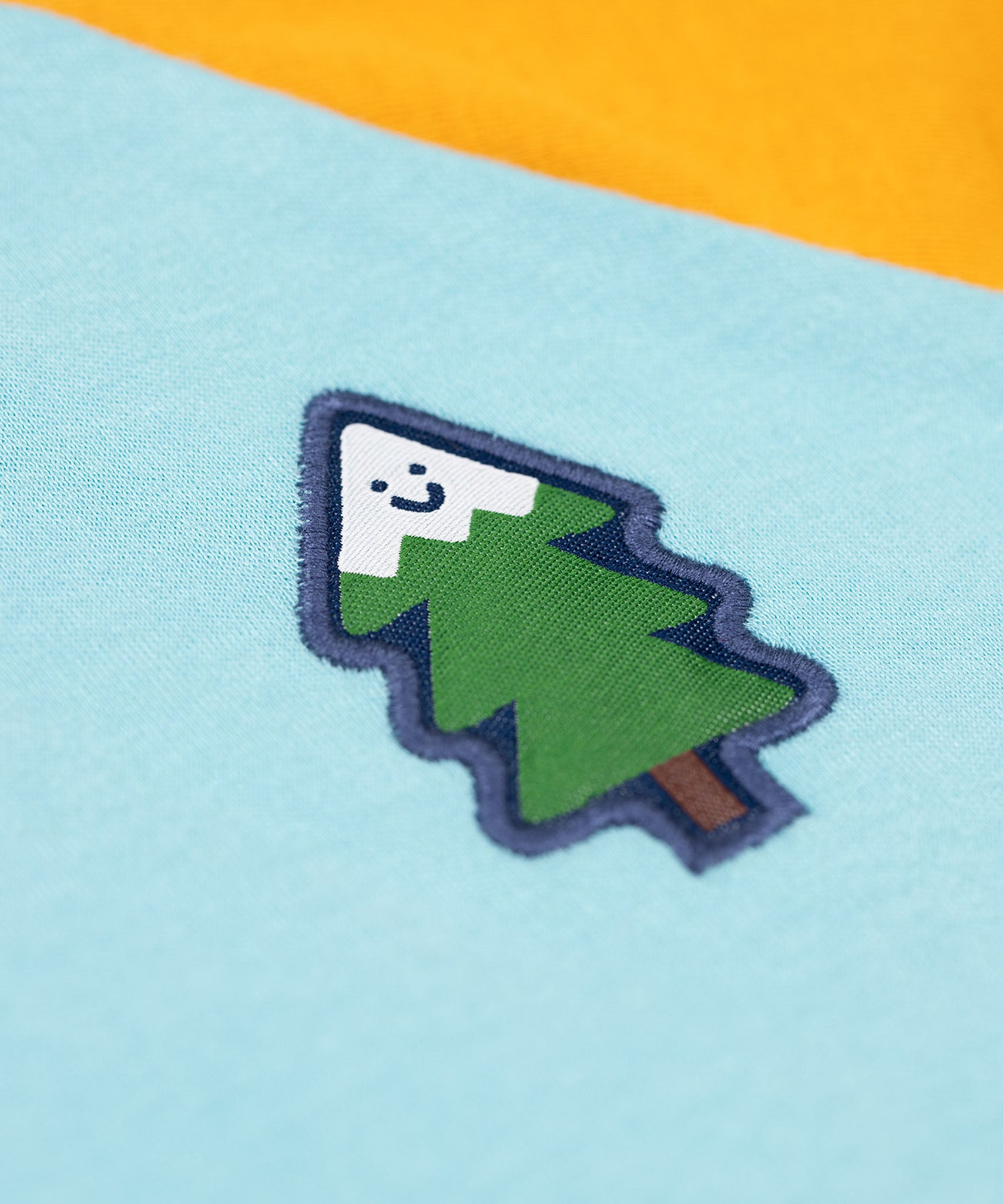 A close up of the small tree detail on the chest of the Frugi Children's Leo raglan long sleeved top. The body of the top is light blue with yellow gold coloured sleeves and a contrasting dark green neck collar. 
