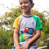 A child sitting on a wooden gate smiling wearing a Frugi raglan t-shirt in a grey marl colour with one green and one yellow sleeve and an adventure themed applique design on the front featuring a pair of green binoculars, camera and a wild card compass. 