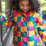 A child wearing the Frugi kids snow and ski coat in rainbow check. A multicoloured check winter coat with 2 side pockets with reflective strips on the arms