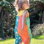 A toddler wearing Frugi Summer Printed Dungaree in the Rainbow Hotchpotch print, featuring the patchwork design and popper fastenings, walking on grass surrounded by plants.