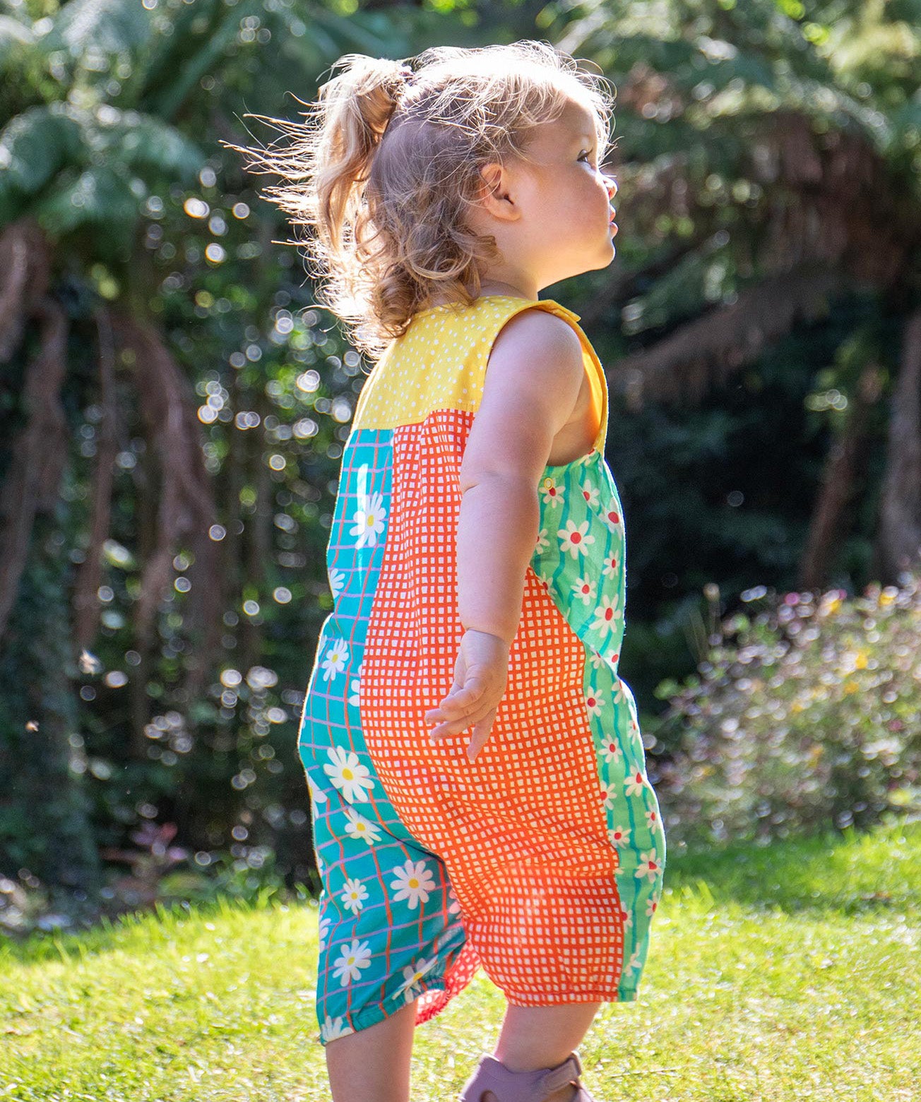 A toddler wearing Frugi Summer Printed Dungaree in the Rainbow Hotchpotch print, featuring the patchwork design and popper fastenings, walking on grass surrounded by plants.