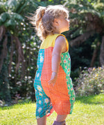A toddler wearing Frugi Summer Printed Dungaree in the Rainbow Hotchpotch print, featuring the patchwork design and popper fastenings, walking on grass surrounded by plants.