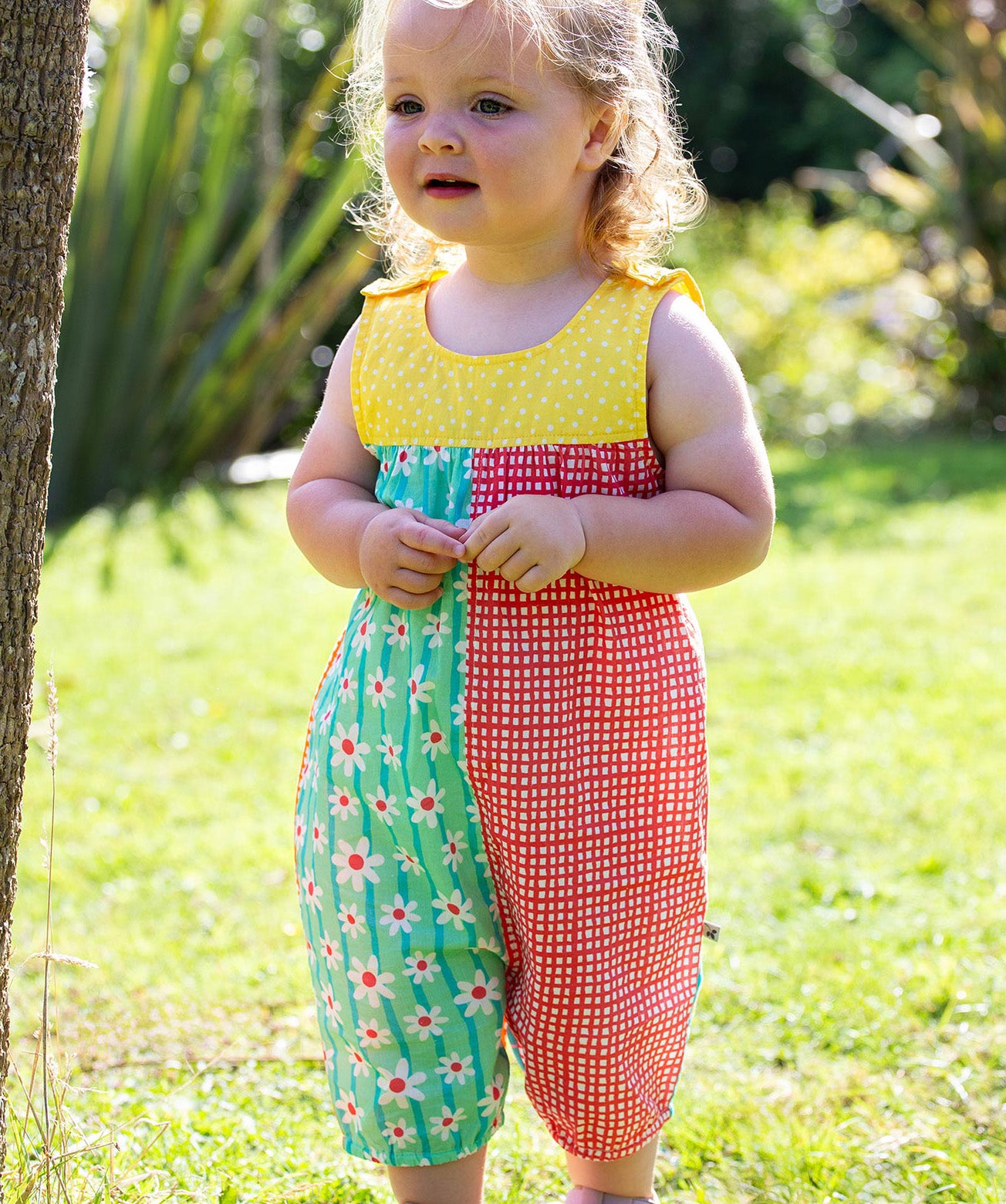 A toddler wearing Frugi Summer Printed Dungaree in the Rainbow Hotchpotch print, featuring the patchwork design and popper fastenings, standing on grass surrounded by plants.