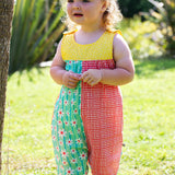 A toddler wearing Frugi Summer Printed Dungaree in the Rainbow Hotchpotch print, featuring the patchwork design and popper fastenings, standing on grass surrounded by plants.