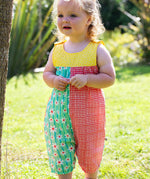 A toddler wearing Frugi Summer Printed Dungaree in the Rainbow Hotchpotch print, featuring the patchwork design and popper fastenings, standing on grass surrounded by plants.