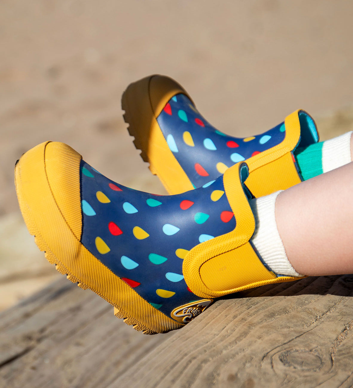 A close up of a child's feet on a wooden surface on a beach wearing Frugi rain or shine rainbow rain design short welly boots with white cotton socks underneath.