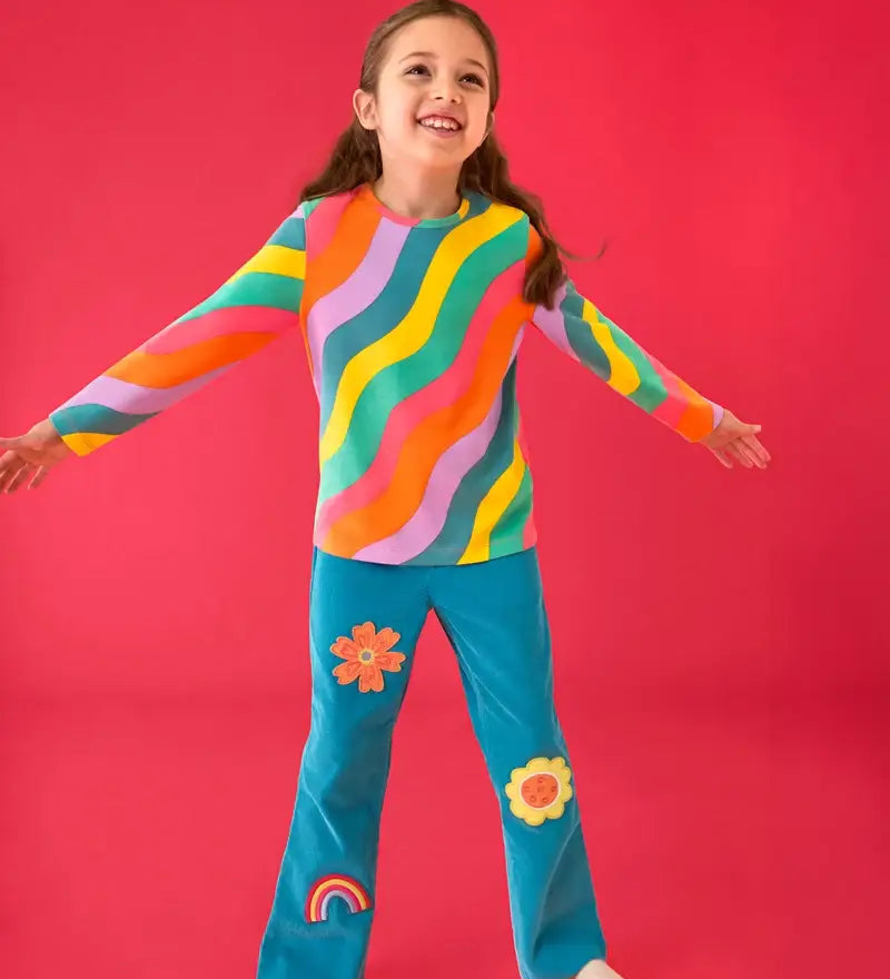 Child wearing the  Frugi rainbow ric rac organic cotton bryher top with the Frugi cherry boat blue corduroy flared trousers. Long sleeved kids top with yellow, blue, lilac, orange, pink and green ric rac stripes. 