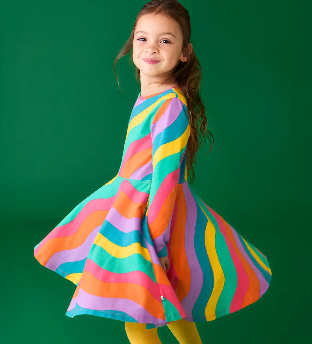 Child twirling wearing the Frugi GOTS organic cotton long sleeved sofia skater dress with a rainbow ric rac pattern.