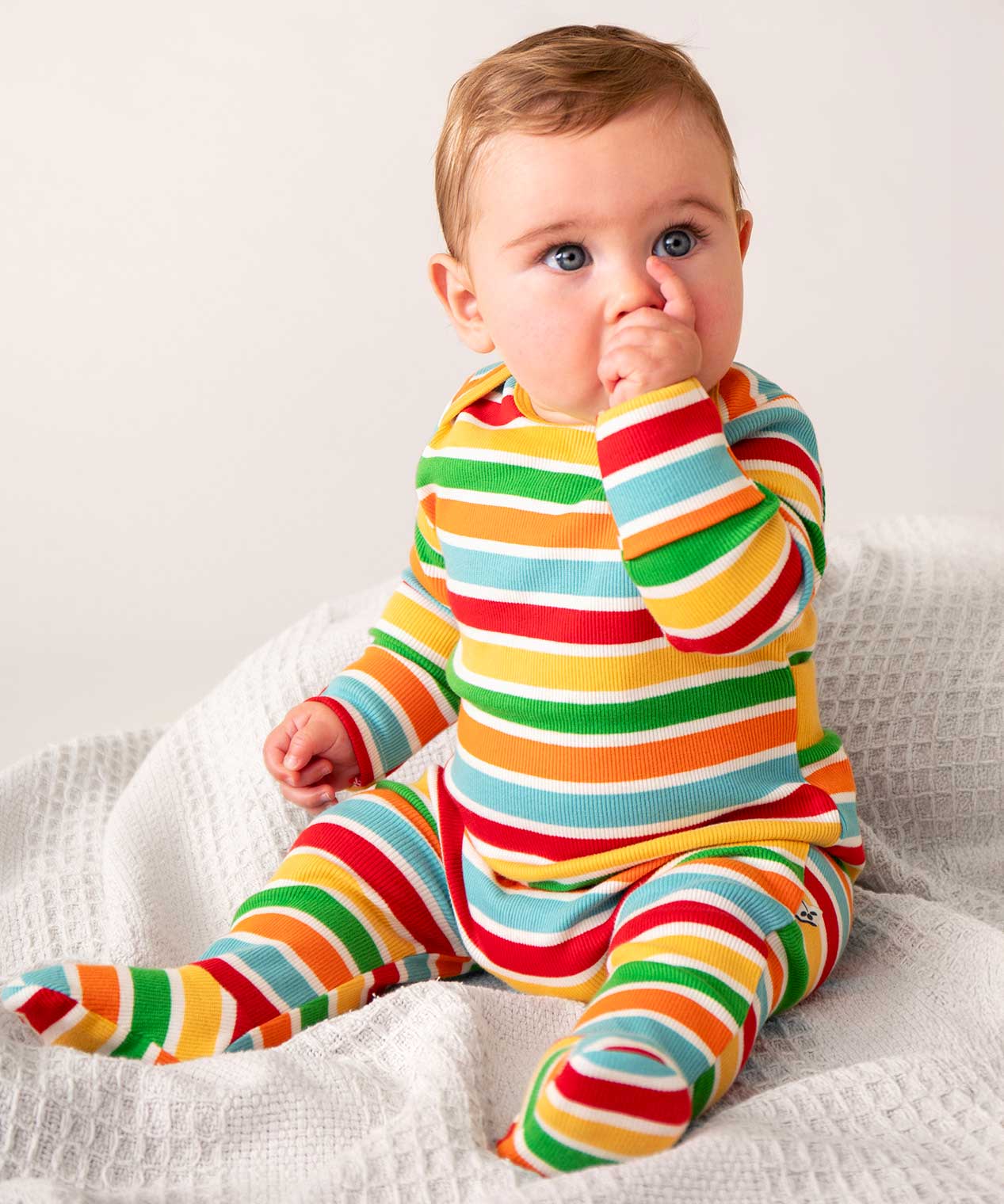 A baby sitting up on white waffle material, sucking their thumb wearing the Frugi easy dressing rainbow striped ribbed material babygrow. 