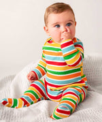 A baby sitting up on white waffle material, sucking their thumb wearing the Frugi easy dressing rainbow striped ribbed material babygrow. 
