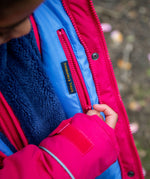 A close up of a child pulling down the pink zip from the hidden side pocket on the inside of the Frugi 3 in 1 snapdragon/ navy  waterproof rambler coat showing the 'treasures & treats' patch stitched on the lilac post consumer recycled polyester fabric