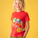 A child standing, in front of a yellow background wearing a Frugi red organic cotton short sleeved kids T-Shirt with a colurful chameleon  on a branch applique on the front with green coloured co-coordinating trousers.