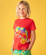 A child standing, in front of a yellow background wearing a Frugi red organic cotton short sleeved kids T-Shirt with a colurful chameleon  on a branch applique on the front with green coloured co-coordinating trousers.