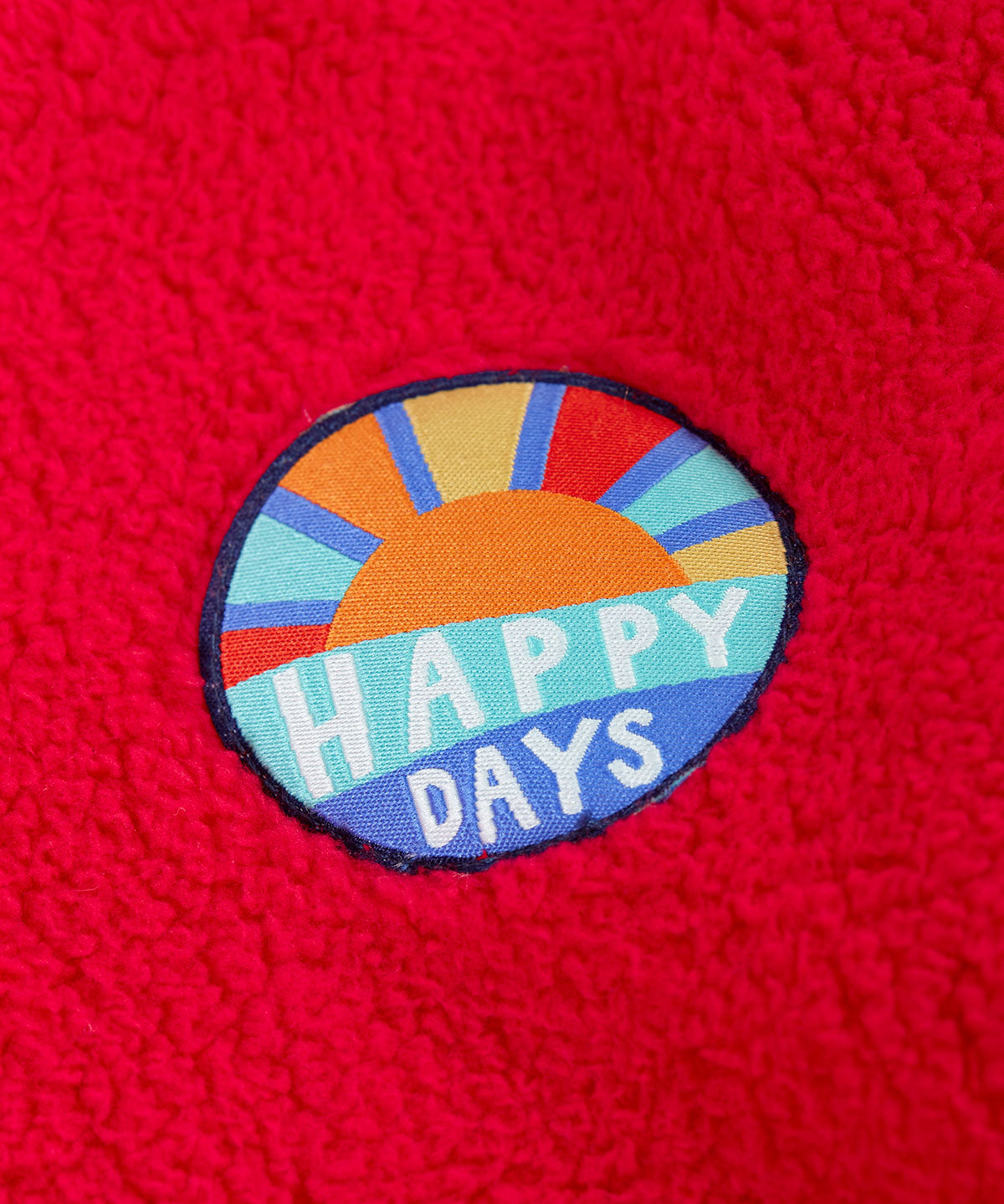A close up of the circle shaped badge on the chest of the Frugi red fleece hooded children's jacket happy days written in white on blue and an image of the sun. The fluffy red fleece ca be seen around it.