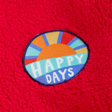 A close up of the circle shaped badge on the chest of the Frugi red fleece hooded children's jacket happy days written in white on blue and an image of the sun. The fluffy red fleece ca be seen around it.