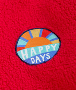 A close up of the circle shaped badge on the chest of the Frugi red fleece hooded children's jacket happy days written in white on blue and an image of the sun. The fluffy red fleece ca be seen around it.